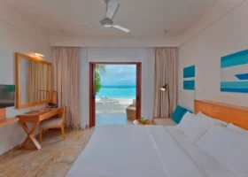 Interno luminoso di una Beach Villa con letto matrimoniale bianco, scrittoio in legno con specchio, ventilatore a soffitto e quadri azzurri alle pareti, porta aperta sulla spiaggia di sabbia bianca con lettini e laguna turchese a Ziyaaraifushi, atollo di Malé Nord, Maldive.