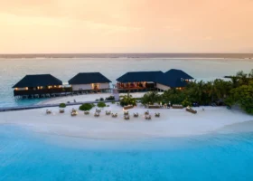Veduta aerea al tramonto di una cena romantica allestita sulla sabbia bianca, con tavoli illuminati da candele davanti ai padiglioni overwater del ristorante dai tetti scuri, laguna turchese e cielo caldo a Ziyaaraifushi, atollo di Malé Nord, Maldive, Oceano Indiano.