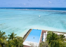 Veduta aerea della piscina a sfioro sul bordo sabbioso con ombrelloni bianchi e palme da cocco, affacciata sulla laguna cristallina con una barca a vela dhoni e la barriera corallina che si infrange all'orizzonte, a Ziyaaraifushi, atollo di Malé Nord, Maldive, Oceano Indiano.