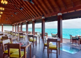 Sala interna del ristorante overwater con alto soffitto in legno a travi a vista, tavoli apparecchiati con cuscini gialli, lati completamente aperti sulla laguna turchese e pannello di vetro sul pavimento affacciato sull'acqua, a Ziyaaraifushi, atollo di Malé Nord, Maldive.