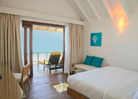 Interno di una Water Villa con letto matrimoniale bianco, soffitto a volta con travi a vista, divanetto con cuscini turchesi e pannello di vetro sul pavimento affacciato sull'acqua; porta scorrevole aperta sulla terrazza con lettino giallo sospesa sulla laguna di Ziyaaraifushi, atollo di Malé Nord, Maldive.