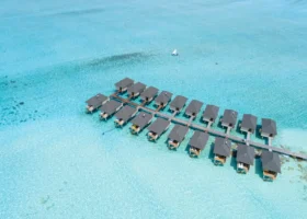 Veduta aerea di due file di ville overwater con tetti scuri disposte lungo il pontile in legno sopra la laguna turchese trasparente, con una barca a vela dhoni maldiviana sullo sfondo, a Ziyaaraifushi, atollo di Malé Nord, Maldive.
