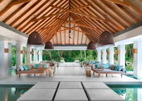 Ampio padiglione open-air con alto tetto a capanna in travi di legno e copertura in strame, lampade a sospensione in rattan intrecciato e ventilatori a soffitto; daybed e divani con cuscini turchesi affacciati su vasche d'acqua a specchio e pedane in pietra, immersi nella vegetazione tropicale di Vakkaru Maldives, Atollo di Baa.