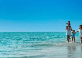Una famiglia con due bambini cammina nell'acqua bassa e cristallina lungo la riva di Vakkaru Maldives: il padre porta il figlio sulle spalle, la madre tiene per mano la bambina; all'orizzonte le ville overwater e il cielo azzurro terso sopra l'Oceano Indiano, Atollo di Baa, Maldive.