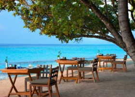 Tavoli e sedie in teak apparecchiati sulla sabbia bianca all'ombra di un grande albero tropicale, lungo la battigia del ristorante Amaany di Vakkaru Maldives, sull'Isola di Vakkaru nell'Atollo di Baa; sullo sfondo la laguna turchese sfuma nell'azzurro dell'Oceano Indiano nella luce tenue del crepuscolo.