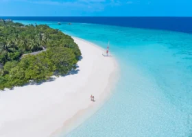 Veduta aerea della spiaggia di sabbia bianca finissima che disegna una curva lungo la laguna dell'Isola di Vakkaru: una coppia passeggia sul bagnasciuga e un catamarano dalla vela colorata è tirato in secca, mentre l'acqua sfuma dal turchese chiaro al blu profondo della barriera corallina, Atollo di Baa, Maldive.