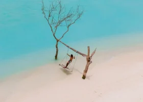 Una persona si dondola su un'altalena di legno con corde sospesa tra due tronchi spogli di legno di deriva, al bordo di una lingua di sabbia bianca, con i piedi quasi a sfiorare l'acqua bassa turchese e brillante dell'isola di Nalaguraidhoo, Atollo di Ari Sud, Maldive, Oceano Indiano.