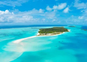 Panoramica aerea dell'intera isola tropicale ricoperta da una fitta foresta di palme da cocco, orlata da bianche lingue di sabbia e da una laguna dalle molteplici sfumature di turchese e blu intenso, con il pontile delle ville overwater sul lato opposto e un cielo azzurro con nuvole sparse, a Nalaguraidhoo, Atollo di Ari Sud, Maldive, Oceano Indiano.