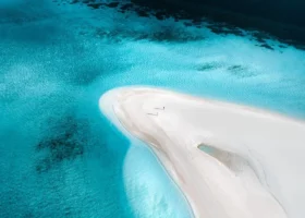 Veduta aerea di una sinuosa lingua di sabbia bianca che emerge tra acque profonde e turchesi, con due minuscole figure che camminano sulla sabbia immacolata e chiazze scure di corallo visibili sotto la superficie cristallina, nei pressi di Nalaguraidhoo, Atollo di Ari Sud, Maldive, Oceano Indiano.