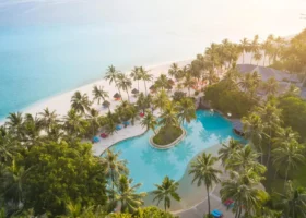 Ripresa aerea di una piscina a forma libera dalle linee sinuose con isolotto centrale di palme, incorniciata da alte palme da cocco e lettini colorati, che si apre su una spiaggia di sabbia bianca e su una laguna turchese immersa nella calda luce dorata del tramonto, presso Nalaguraidhoo, Atollo di Ari Sud, Maldive.