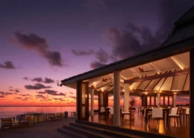 Ristorante overwater a pianta aperta con tetto spiovente, colonne e travi in legno, ventilatori a soffitto e tavoli apparecchiati con tovagliato bianco, affacciato su un pavimento in legno con poltroncine lounge, mentre un cielo al tramonto in intense tonalità viola e arancio si riflette sull'oceano calmo a Nalaguraidhoo, Atollo di Ari Sud, Maldive.
